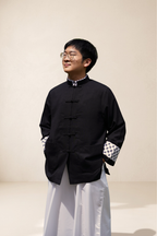 A man wearing a black traditional Chinese-style jacket with white pants, standing against a plain light gray background.