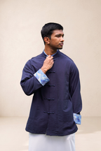 A man wearing a navy blue traditional-style jacket with blue patterned cuffs and a light-colored kurta, standing against a plain beige background.