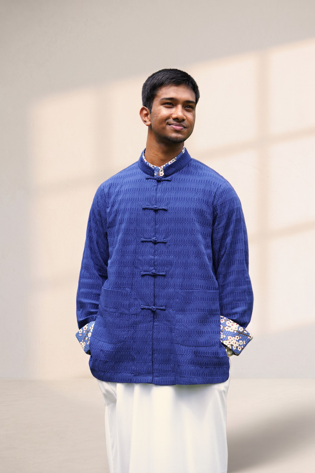 A man wearing a textured royal blue traditional Chinese-style jacket with dark buttons and patterned cuffs, paired with white trousers, standing against a solid gray background.