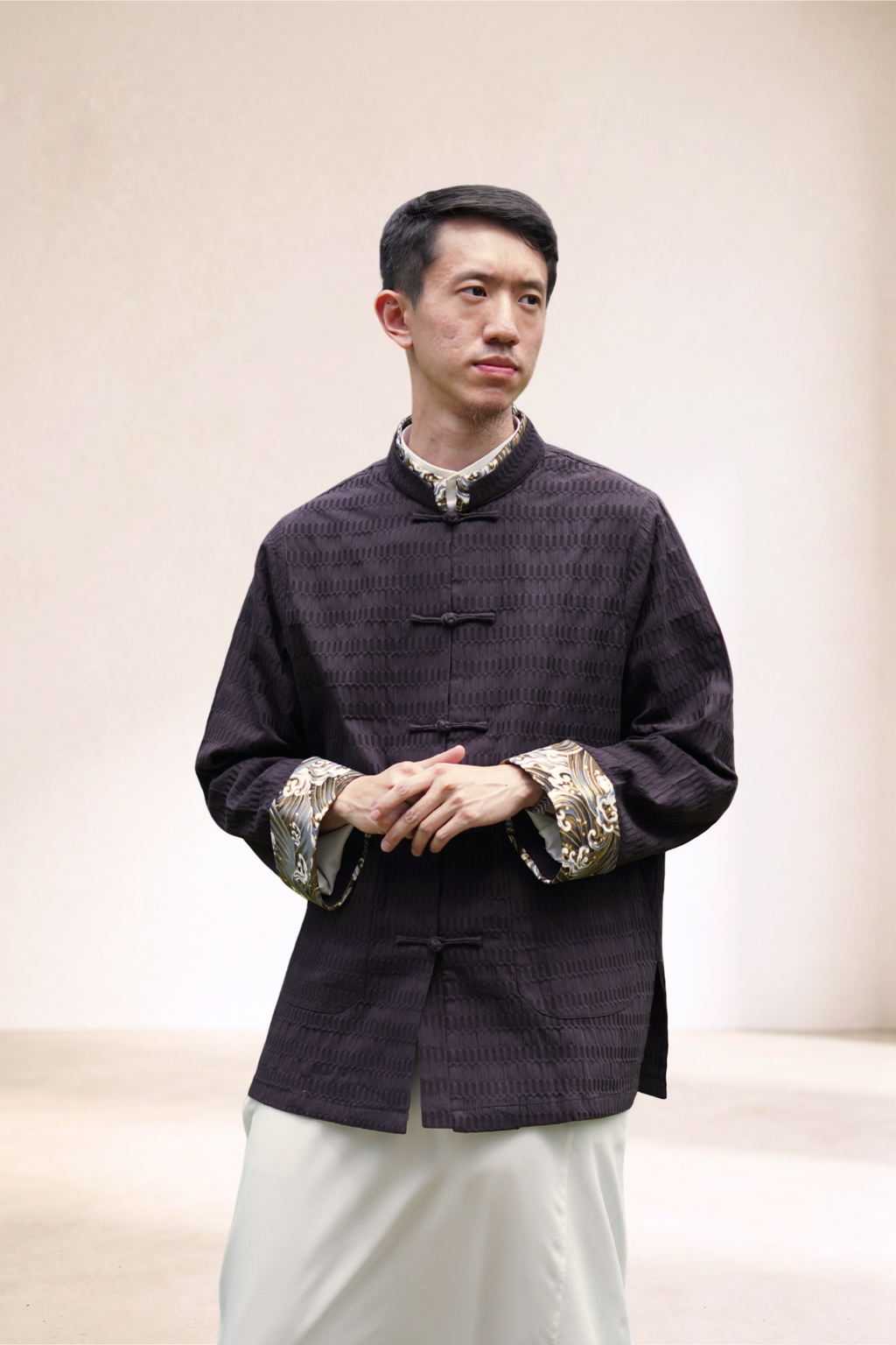 A man wearing a dark textured traditional Chinese-style jacket with ornate gold patterned cuffs, paired with white pants, standing against a solid gray studio background.